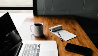На столе стоит ноутбук, кофе, блокнот с ручкой и смартфон